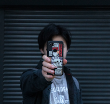 A person holds a smartphone in front of their face, with the phone's case covered in multiple stickers featuring various designs and text. The background consists of a dark, ribbed surface.