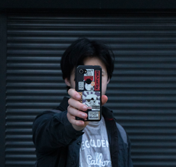 A person holds a smartphone in front of their face, with the phone's case covered in multiple stickers featuring various designs and text. The background consists of a dark, ribbed surface.
