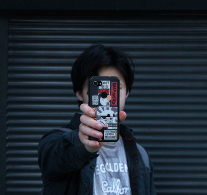 A person holds a smartphone in front of their face, with the phone's case covered in multiple stickers featuring various designs and text. The background consists of a dark, ribbed surface.