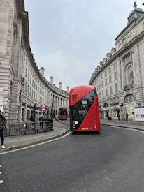 A red double-decker bus is driving along a curved city street bordered by elegant, historic buildings with classical architecture. The buildings feature large windows and detailed stone facades. There is a 'London Underground' sign visible near a group of pedestrians on the sidewalk.