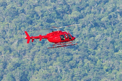 A red helicopter is flying over a dense forest landscape, with trees stretching far into the distance. The helicopter appears to be mid-flight with spinning rotors, contrasting against the lush green foliage below.