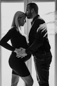 A black and white image depicts a couple standing close together in front of tall windows. The woman, who appears to be pregnant, is wearing a fitted dress, and is gently holding her belly. The man, wearing a shirt and pants, leans in to kiss her forehead. Light streams in through the windows, silhouetting their figures and creating a serene atmosphere.