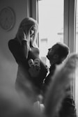 A black and white image featuring a pregnant woman standing near a window with a man kneeling in front of her, holding a pair of baby shoes against her belly. The woman smiles gently at the man, capturing a tender and intimate moment. A wall clock is visible in the background, adding a touch of domesticity to the scene.