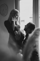 A black and white image featuring a pregnant woman standing near a window with a man kneeling in front of her, holding a pair of baby shoes against her belly. The woman smiles gently at the man, capturing a tender and intimate moment. A wall clock is visible in the background, adding a touch of domesticity to the scene.
