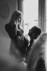A black and white image featuring a pregnant woman standing near a window with a man kneeling in front of her, holding a pair of baby shoes against her belly. The woman smiles gently at the man, capturing a tender and intimate moment. A wall clock is visible in the background, adding a touch of domesticity to the scene.