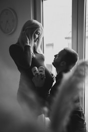 A black and white image featuring a pregnant woman standing near a window with a man kneeling in front of her, holding a pair of baby shoes against her belly. The woman smiles gently at the man, capturing a tender and intimate moment. A wall clock is visible in the background, adding a touch of domesticity to the scene.