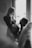 A black and white image featuring a pregnant woman standing near a window with a man kneeling in front of her, holding a pair of baby shoes against her belly. The woman smiles gently at the man, capturing a tender and intimate moment. A wall clock is visible in the background, adding a touch of domesticity to the scene.