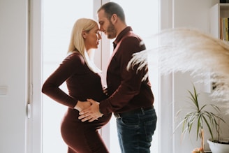 A couple stands closely in an intimate, tender moment by a window. The light softly filters through, casting a warm glow around them. The woman has blonde hair and is wearing a fitted maroon dress, showcasing her pregnant belly, while the man wears a matching maroon shirt and dark jeans. They gently rest their hands on her belly, symbolizing affection and anticipation of their child.