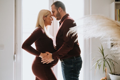 A couple stands closely in an intimate, tender moment by a window. The light softly filters through, casting a warm glow around them. The woman has blonde hair and is wearing a fitted maroon dress, showcasing her pregnant belly, while the man wears a matching maroon shirt and dark jeans. They gently rest their hands on her belly, symbolizing affection and anticipation of their child.
