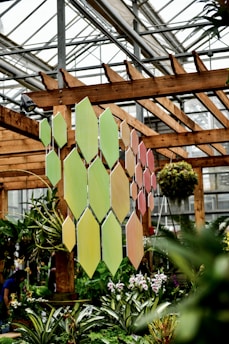 A greenhouse features vibrant green plants and colorful orchids. A decorative panel composed of diamond-shaped tiles in shades of green and pink hangs from a wooden structure. The greenhouse has a metal and glass ceiling allowing natural light to illuminate the plants.