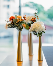 Three modern, tall, metallic vases are arranged on a light-colored table. Each vase holds a vibrant bouquet composed of white hydrangeas, orange and yellow flowers, and green foliage. The background is softly blurred, indicating an indoor setting with natural light streaming through large windows.