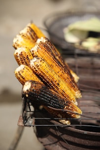 Charred ears of corn are stacked on a metal grill, showcasing a mix of golden yellow and dark brown colors from being roasted. The grill itself appears rustic and is likely part of a street food setup.