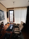 A modern home office setup featuring a sleek wooden desk equipped with a curved monitor, keyboard, and mouse. A comfortable, brown leather office chair is positioned in front of the desk. The room is well-lit by natural light coming through a large window with light-colored curtains. A painting of Albert Einstein is mounted on the wall beside the window. The decor is minimalist and clean.