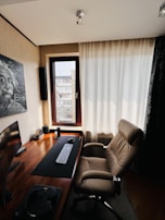 A modern home office setup featuring a sleek wooden desk equipped with a curved monitor, keyboard, and mouse. A comfortable, brown leather office chair is positioned in front of the desk. The room is well-lit by natural light coming through a large window with light-colored curtains. A painting of Albert Einstein is mounted on the wall beside the window. The decor is minimalist and clean.