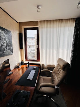 A modern home office setup featuring a sleek wooden desk equipped with a curved monitor, keyboard, and mouse. A comfortable, brown leather office chair is positioned in front of the desk. The room is well-lit by natural light coming through a large window with light-colored curtains. A painting of Albert Einstein is mounted on the wall beside the window. The decor is minimalist and clean.