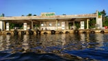A fuel station located along a waterfront, with multiple fuel dispensers visible. The building appears weathered, with visible wear and rust, and is surrounded by lush greenery in the background. The calm water reflects the station and the blue sky above, creating a serene atmosphere.