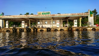 A fuel station located along a waterfront, with multiple fuel dispensers visible. The building appears weathered, with visible wear and rust, and is surrounded by lush greenery in the background. The calm water reflects the station and the blue sky above, creating a serene atmosphere.