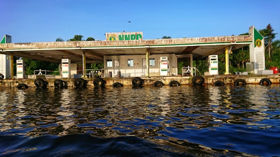 A fuel station located along a waterfront, with multiple fuel dispensers visible. The building appears weathered, with visible wear and rust, and is surrounded by lush greenery in the background. The calm water reflects the station and the blue sky above, creating a serene atmosphere.