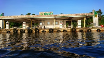 A fuel station located along a waterfront, with multiple fuel dispensers visible. The building appears weathered, with visible wear and rust, and is surrounded by lush greenery in the background. The calm water reflects the station and the blue sky above, creating a serene atmosphere.