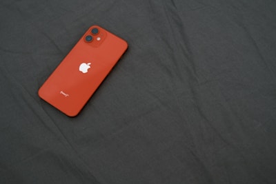 A red smartphone with a white apple logo placed on a dark fabric surface. The phone has dual cameras on the top left corner.