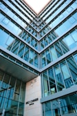 A modern architectural building with multiple glass windows reflecting the sky, presented from a low-angle perspective. The structure features a geometric design, and there's signage indicating 'Váci Corner Offices' near the entrance.