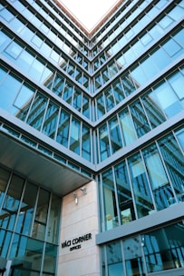 A modern architectural building with multiple glass windows reflecting the sky, presented from a low-angle perspective. The structure features a geometric design, and there's signage indicating 'Váci Corner Offices' near the entrance.