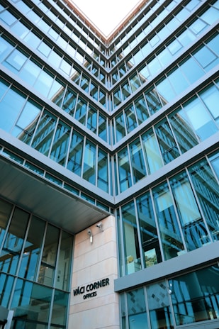 A modern architectural building with multiple glass windows reflecting the sky, presented from a low-angle perspective. The structure features a geometric design, and there's signage indicating 'Váci Corner Offices' near the entrance.