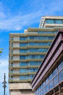 A modern multi-story building with large glass windows and a concrete structure is set against a bright blue sky. The architecture features clean lines and a minimalist design. A dark metal street lamp is visible on the left side, and part of another building with reflective glass panels is in the foreground.