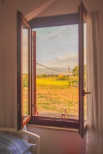 An open window reveals a scenic view of a countryside landscape with green fields, trees, and hills in the distance. The window frame is wooden, and a partial view of an indoor room with a bed is visible. The sky is blue with some clouds, casting a warm, soothing glow.