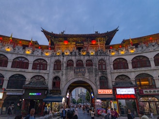 A traditional Chinese building with multiple arched windows, adorned with vibrant red lanterns and flags. The building features intricate roof decorations and appears to be a busy marketplace with a variety of shops, as people are milling about in the area.