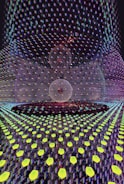 A visually complex scene featuring a symmetrical pattern with a central spherical object overlaid with a grid of glowing hexagonal shapes. The spherical object appears to be composed of intersecting lines, creating a wireframe look. The background is filled with a vibrant array of colors and reflections, giving a sense of depth and digital texture.
