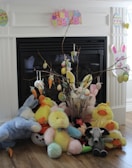 A festive Easter display in front of a fireplace features plush toys including bunnies and ducks, a decorative branch with hanging eggs and carrots, and colorful 'Happy Easter' signage. The setting suggests a cheerful and playful atmosphere suitable for Easter celebrations.