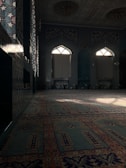 A richly decorated interior with intricate tile patterns covering the walls and archways. Light filters in through arched windows, casting shadows across a carpeted floor adorned with elaborate designs. The overall feel is one of elegance and ornate craftsmanship, typical of traditional Islamic architecture.