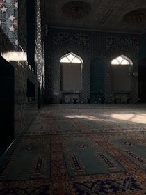 A richly decorated interior with intricate tile patterns covering the walls and archways. Light filters in through arched windows, casting shadows across a carpeted floor adorned with elaborate designs. The overall feel is one of elegance and ornate craftsmanship, typical of traditional Islamic architecture.