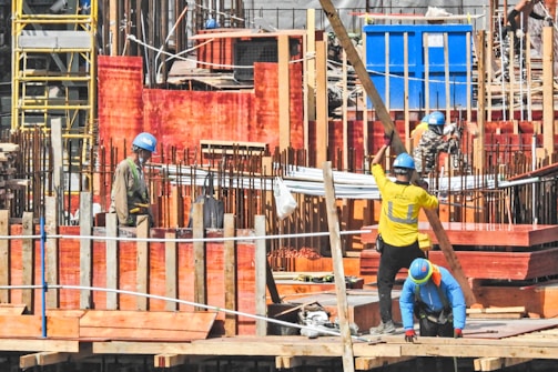 A busy construction site with several workers wearing safety helmets and reflective vests. They are surrounded by scaffolding, wooden planks, and stacks of bricks. Metal rods are protruding from various structures, and a blue container is visible in the background.