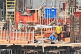 Several construction workers wearing blue helmets and safety vests are actively engaged on a construction site. The background is filled with scaffolding, red wooden planks, and metal rods, indicating the structural framework of an ongoing building project.