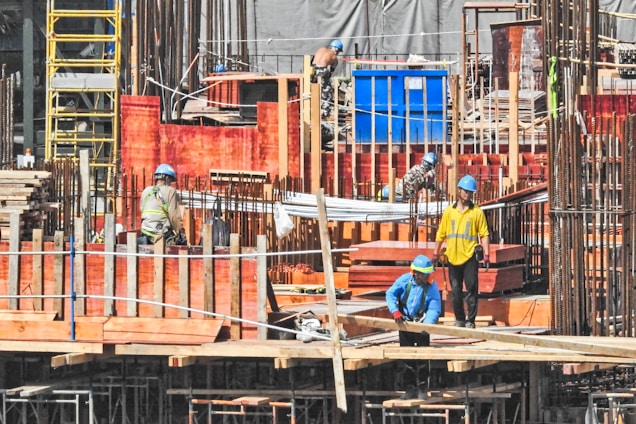 Several construction workers wearing blue helmets and safety vests are actively engaged on a construction site. The background is filled with scaffolding, red wooden planks, and metal rods, indicating the structural framework of an ongoing building project.