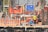 Several construction workers wearing blue helmets and safety vests are actively engaged on a construction site. The background is filled with scaffolding, red wooden planks, and metal rods, indicating the structural framework of an ongoing building project.
