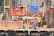 Several construction workers wearing blue helmets and safety vests are actively engaged on a construction site. The background is filled with scaffolding, red wooden planks, and metal rods, indicating the structural framework of an ongoing building project.