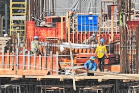 Several construction workers wearing blue helmets and safety vests are actively engaged on a construction site. The background is filled with scaffolding, red wooden planks, and metal rods, indicating the structural framework of an ongoing building project.