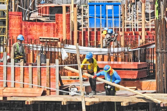 Construction site bustling with several workers wearing safety gear including helmets and high-visibility clothing. The site is structured with wooden formwork and iron rods, indicating active building activity. Workers are handling materials and tools, suggesting an ongoing construction process.