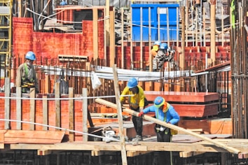 Construction site bustling with several workers wearing safety gear including helmets and high-visibility clothing. The site is structured with wooden formwork and iron rods, indicating active building activity. Workers are handling materials and tools, suggesting an ongoing construction process.