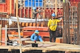 Three construction workers wearing safety gear and hard hats are actively engaged in building activities at a construction site. They are surrounded by metal rebar, wooden planks, and other construction materials.