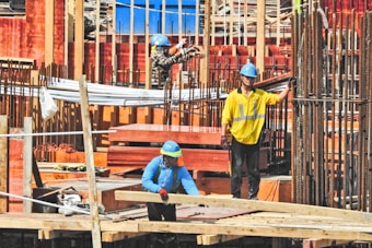 Three construction workers wearing safety gear and hard hats are actively engaged in building activities at a construction site. They are surrounded by metal rebar, wooden planks, and other construction materials.