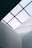 A minimalist architectural perspective captures geometric lines and angular shapes of a building's interior, highlighted by the absence of colors that emphasizes structure. A glass ceiling with a grid pattern allows diffused natural light to spill into the space, casting soft shadows.
