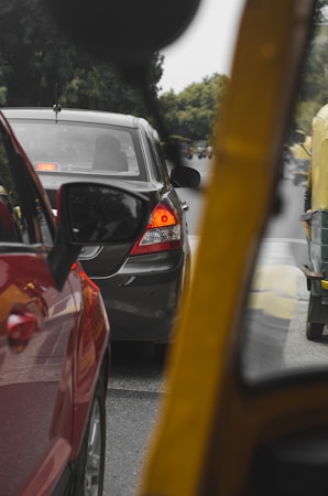 Cars are closely lined up in traffic on a road surrounded by greenery, with a focus on a dark-colored sedan and hints of other vehicles, including a red car and a yellow auto-rickshaw.