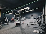 A modern gym interior equipped with various fitness machines such as treadmills, rowing machines, and a punching bag. The room has a dark color scheme with green accent lighting running across the ceiling and walls. The floor is carpeted, and there's a sense of tidy organization.