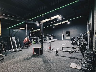 A modern gym interior equipped with various fitness machines such as treadmills, rowing machines, and a punching bag. The room has a dark color scheme with green accent lighting running across the ceiling and walls. The floor is carpeted, and there's a sense of tidy organization.