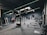 A modern gym interior equipped with various fitness machines such as treadmills, rowing machines, and a punching bag. The room has a dark color scheme with green accent lighting running across the ceiling and walls. The floor is carpeted, and there's a sense of tidy organization.