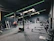 A modern gym interior equipped with various fitness machines such as treadmills, rowing machines, and a punching bag. The room has a dark color scheme with green accent lighting running across the ceiling and walls. The floor is carpeted, and there's a sense of tidy organization.